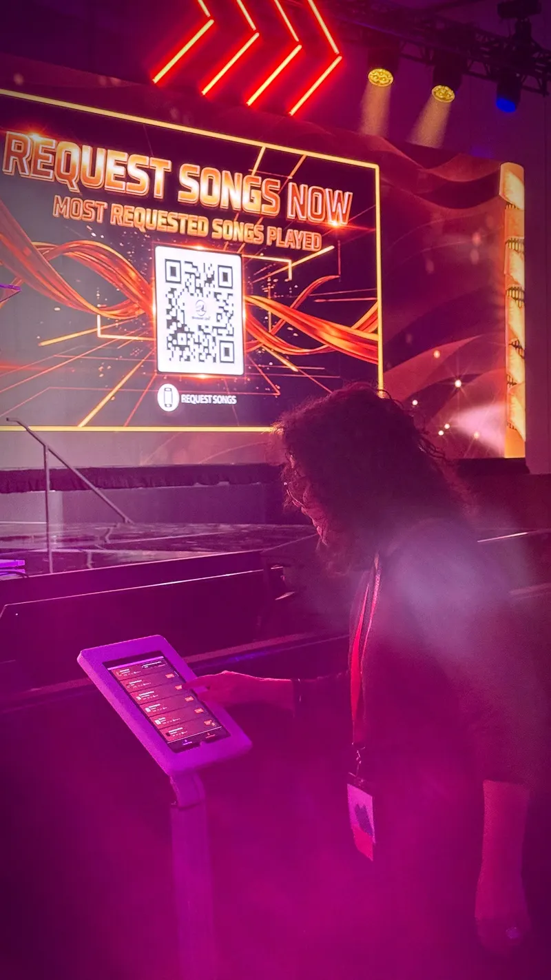 A guest choosing a song on a uRequest Live touchscreen kiosk beneath a REQUEST SONGS NOW screen at a live event