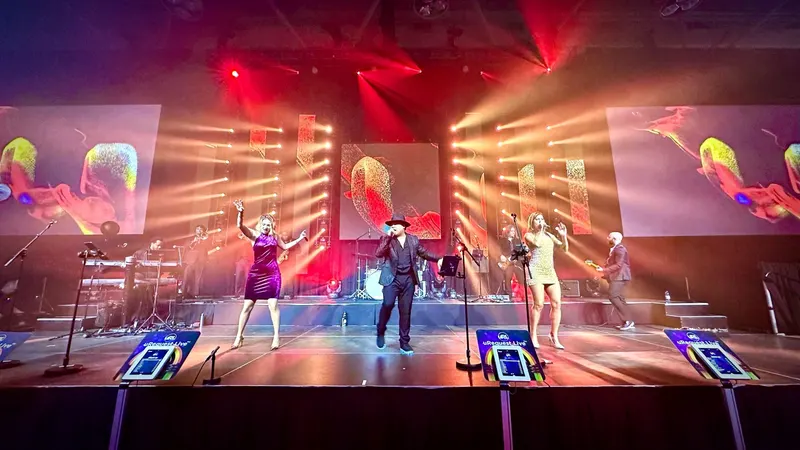 Three uRequest Live vocalists on a red and amber production stage with song-request kiosks along the stage edge
