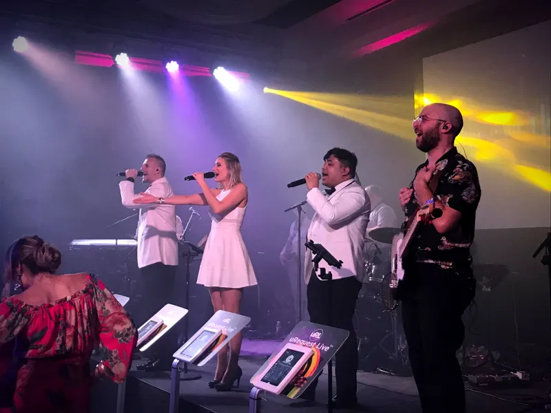 Four vocalists dressed in white with request kiosks while a guest dances in the foreground